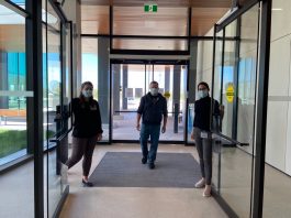 Deux femmes qui ouvrent les portes d'un édifice vitré et un homme qui entre. Tous masqués.