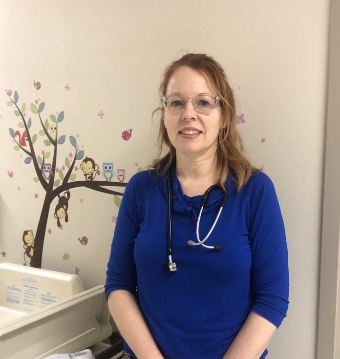 Cathy Cathy, vêtue d'un uniforme bleu, et portant un stéthoscope sur l'épaule, souriante dans un corridor de l'hôpital
