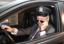 Dr Leduc déguisé en chauffeur de limousine, le logo d'Agrément sur sa casquette. Il a baissé ses lunettes de soleil et fait un clin d'oeil.